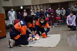 女性消防団員3人が乳児の救命処置デモンストレーションをする写真　周りで参加者が見ている様子