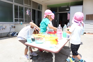 テーブルに並べられたペットボトルの中に色水が入っている。子ども達がテーブルの周りで遊んでいる