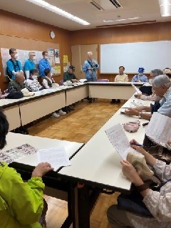 ロの字に囲まれた机で、史跡ガイドの会の方の説明を聞く参加者の写真
