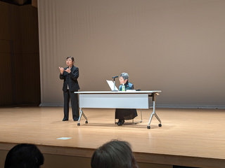 舞台の上で辻野さんが話す様子の写真。机といすが用意されており、椅子に座った状態で話している。隣には手話通訳もいる。