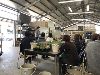 鉢植えが並ぶ机で参加者が講師の話を聞いている様子の写真
