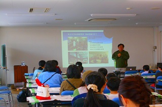 講師がプロジェクターで映された資料の前で話す様子の写真。多数の参加者が講師を見て話を聞いている。