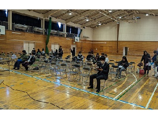 体育館にパイプ椅子が並べられ、何人かが座っている様子の写真