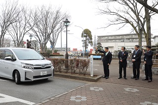 出発する車を見送る井崎市長・副市長を含めた職員4人の写真。市長の横には「流山市・能登町　姉妹都市締結記念樹」の表札がある。