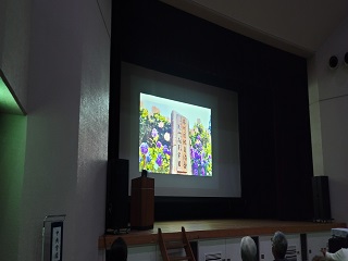 舞台上のスクリーンに木の札と花の写真が投影されている様子の写真