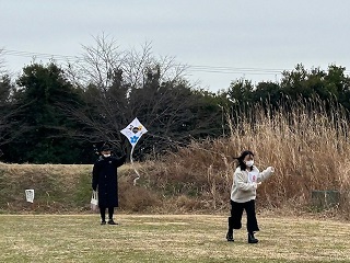 芝の上で子どもがたこの糸を持ち、走っていく様子の写真。奥の男性がたこを持っている。