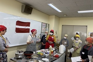 外国人の講師が調理室でエプロンをつけている市民に対して講義を行っている様子の写真
