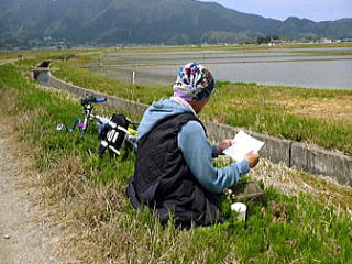 田んぼのあぜ道で視聴者の手紙を読む火野さん